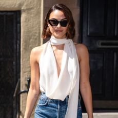 Camilla Mendes steps outside wearing stovepipe jeans with a scarf blouse and white heeled sandals.