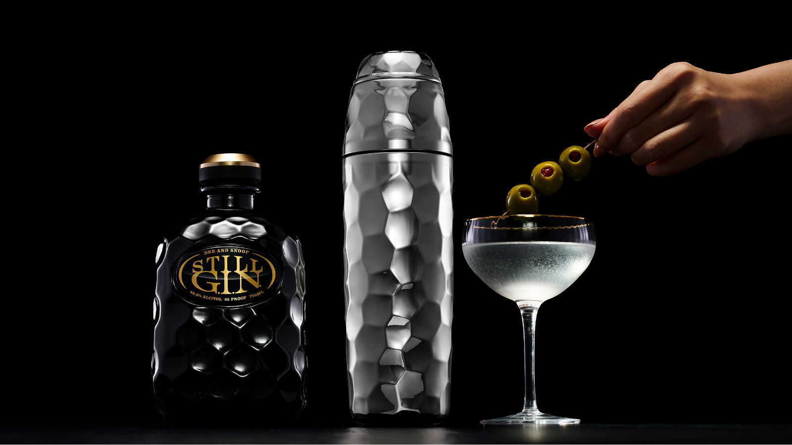 Hammered silver cocktail shaker next to martini glass and black gin bottle