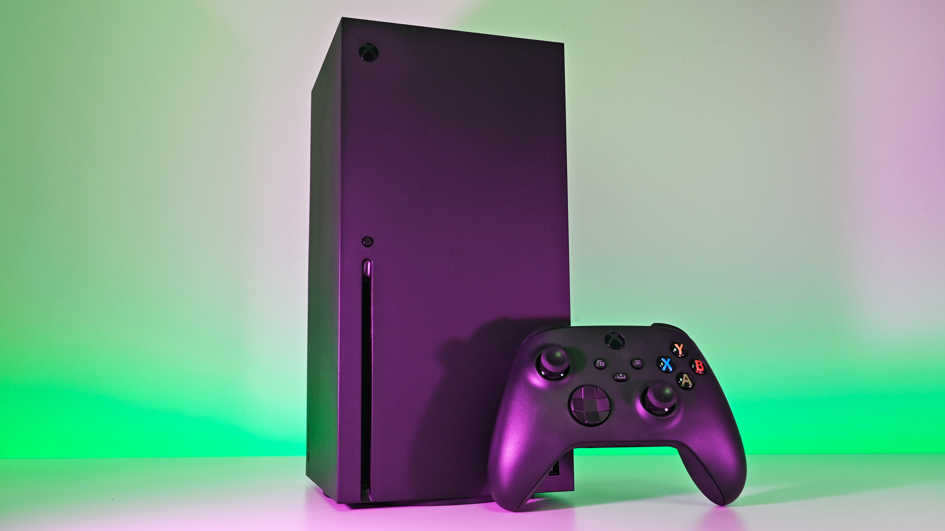 Vertical black Xbox Series X console and controller sitting together, dramatized by vibrant green and purple background lights.