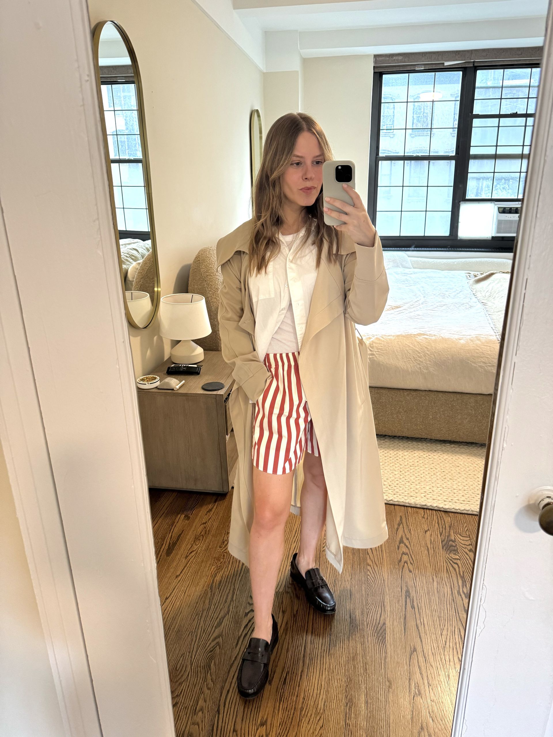 Nikki Chwatt wears red striped shorts, a white button-down shirt, a white t-shirt, burgundy loafers, and a beige trench coat.