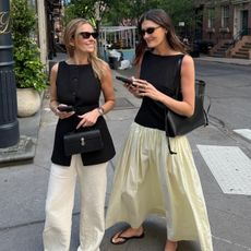 NYC style influencer Coco Schiffer with a friend walking at a crosswalk both wearing stylish spring outfit ideas with black sunglasses, a two-tone dress, black vest, black bags, and flip-flops