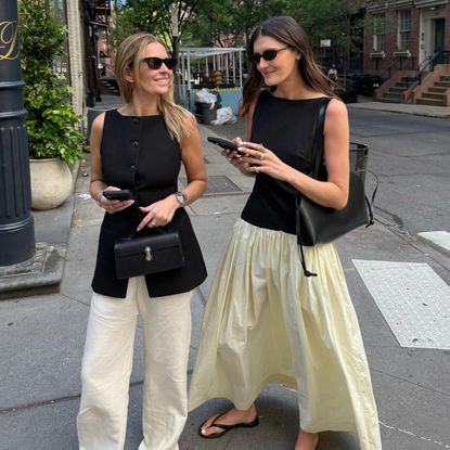 NYC style influencer Coco Schiffer with a friend walking at a crosswalk both wearing stylish spring outfit ideas with black sunglasses, a two-tone dress, black vest, black bags, and flip-flops