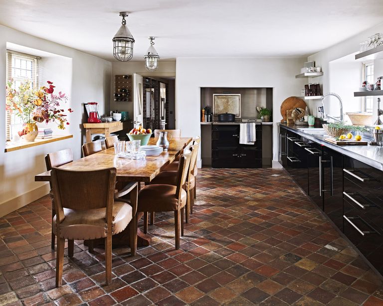 Kitchen flooring ideas. Kitchen diner with dark units, grey walls, terracotta tiled floor and wooden table and chairs. Stainless steel work surface and open shelves.