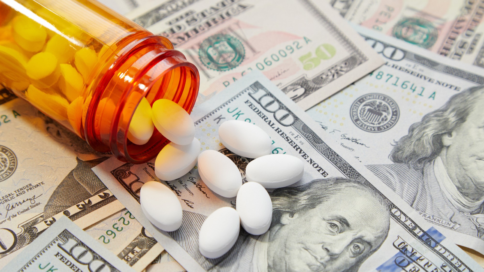 A close-up shot of a spilled amber prescription pill bottle with several white, oval pills resting on a background of various US dollar bills