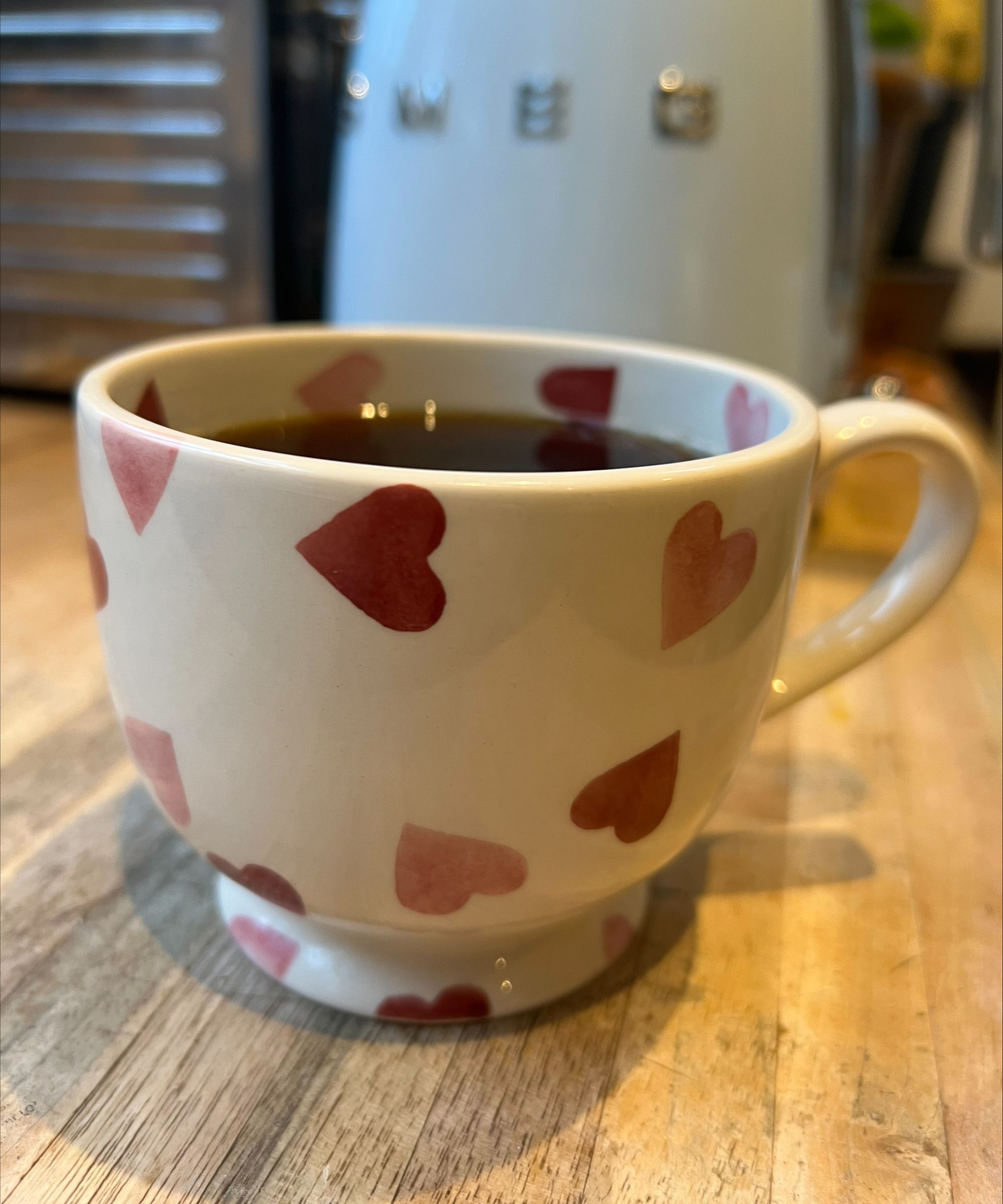 A cup of black coffee in a white cup with pink hearts on standing on a wooden countertop, made with the CHEMEX coffee maker. In the background is the side of a silver Dualit toaster, and a baby blue SMEG kettle seen out of focus.