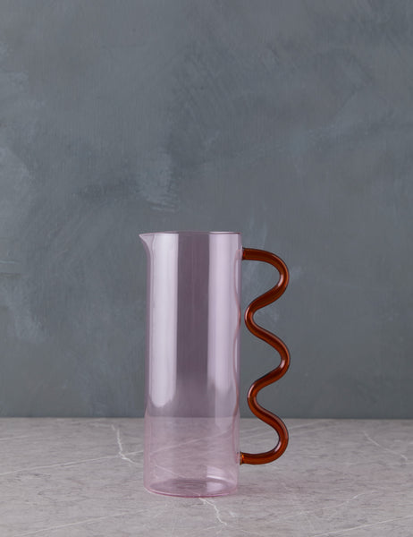 Lulu and Georgia pink and orange glass wave Pitcher sitting on top of marble countertop, dark gray background.