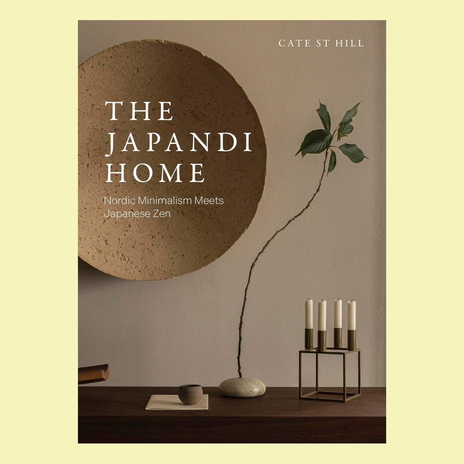 A hardcover book with a snapshot of a wood surface with a leafy stem on a pebble base, a candlestick holder, and a mini ceramic bowl by a textured wall-mounted plate. The book is titled 'The Japandi Home: Nordic Minimalism Meets Japanese Zen' by Cate St Hill.