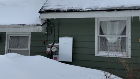 A Tesla Powerwall next to a utility meter on the exterior wall of a green house