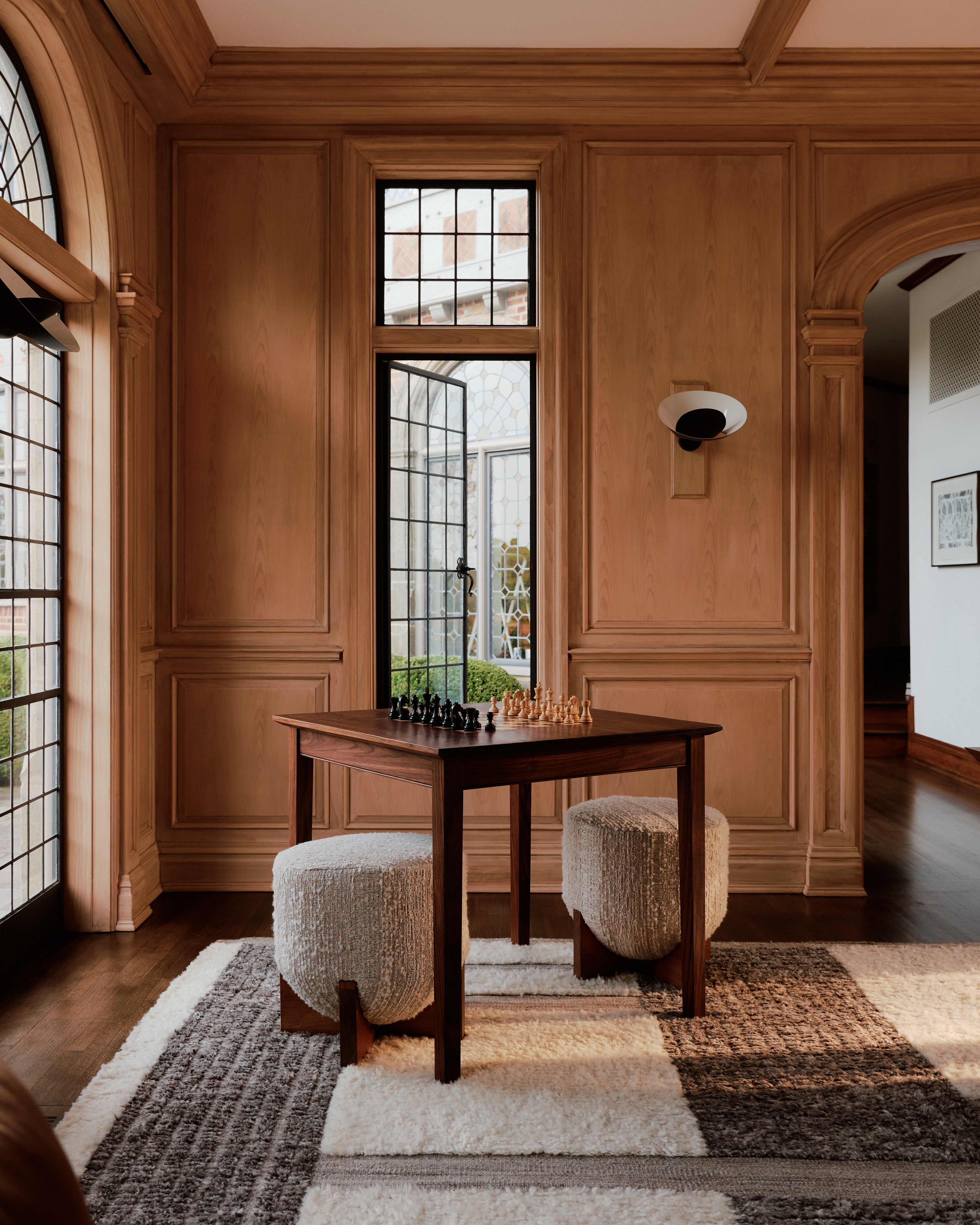 A wooden table styled with two gray stools sits in a corner of a grand room with detailed wooden walls, mosaic floors, and arched windows.