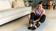 woman ready to exercise in a living room on an exercise mat strapping ankle weights around her ankles. 