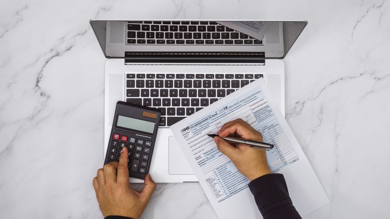 A person&#039;s hand holds a pen above a tax form in front of an open laptop and next to a calculator.