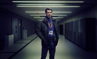 Kelvin Fletcher as Jack, standing in a school corridor wearing a blue shirt, blue trousers, a striped tie, a green parka and a staff ID laminate, with his hands in his jacket pockets