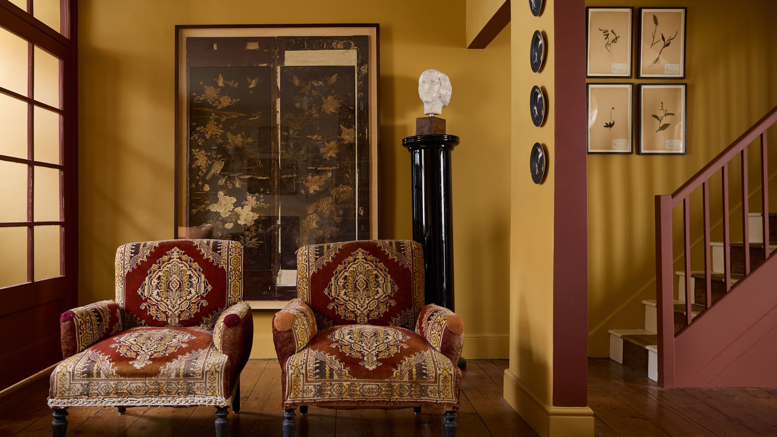 Living room with mustard yellow walls, a burgundy staircase and door frames, a large framed antique picture, two red tapestry armchairs, a plinth with a bust on top, and a wall of botanical prints.
