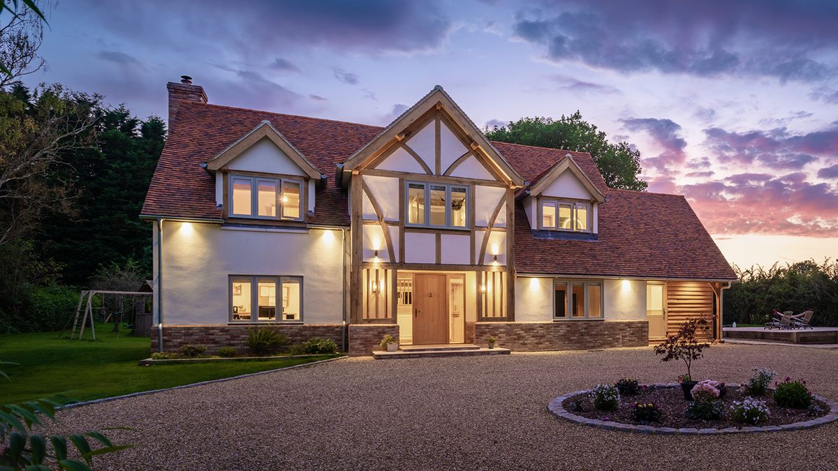 A beautiful two-storey oak frame home at twilight with exterior lights glowing. There is a large gravel driveway at the front of the house