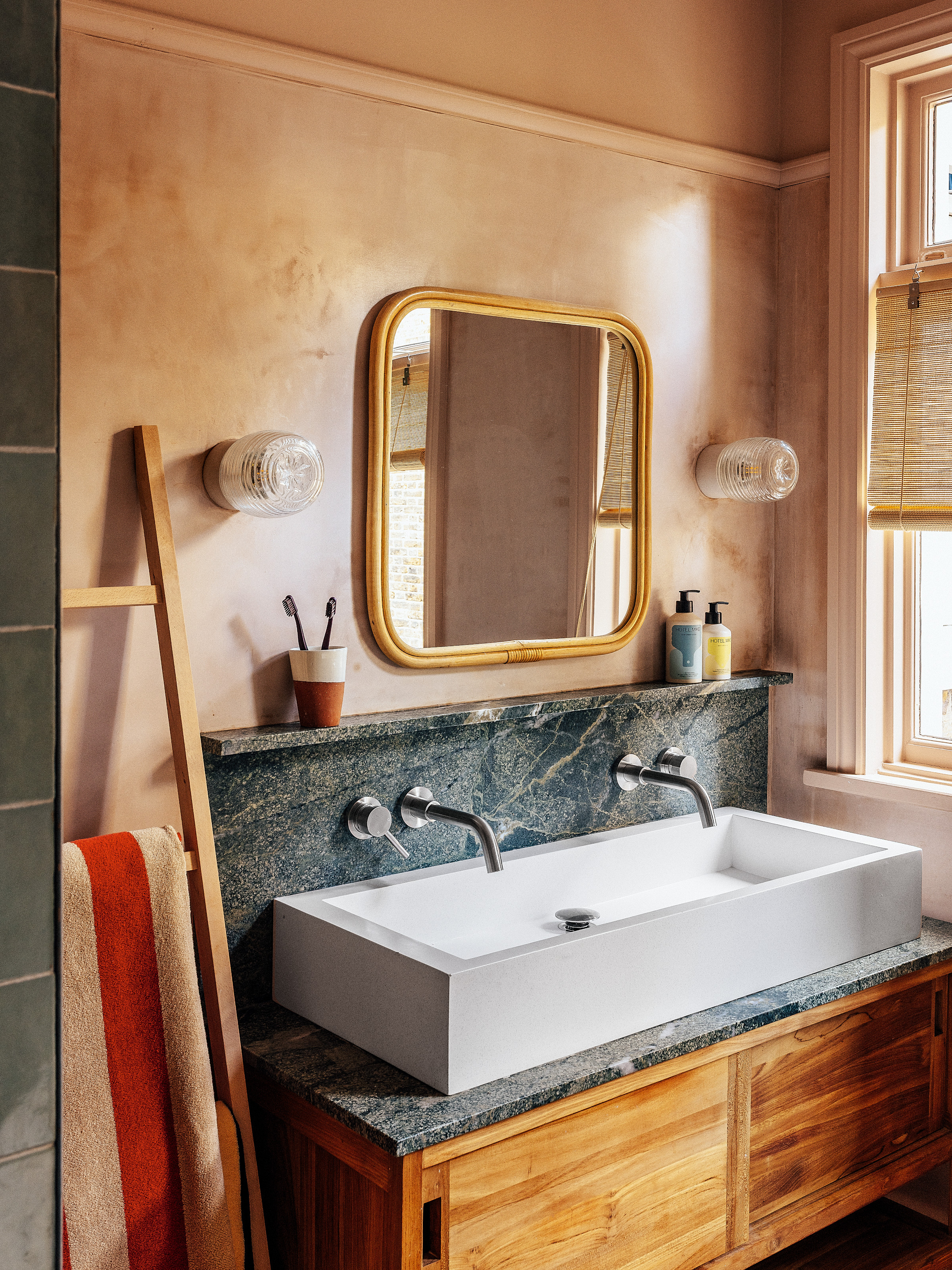 Bathroom with plaster pink walls, wood vanity with double sink and blue stone countertop, square bamboo mirror and pink and red striped towel