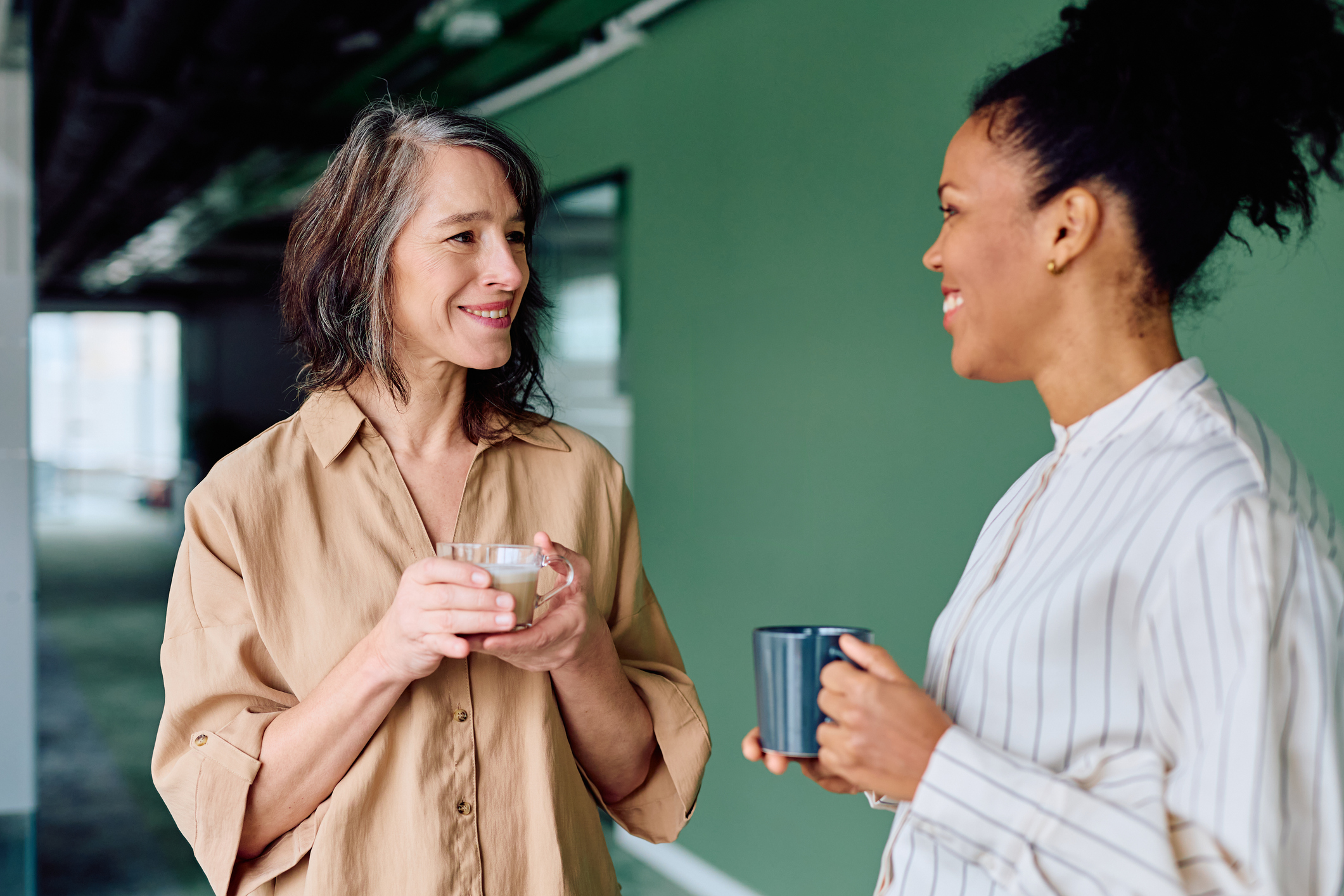 Two diverse women colleagues smiling and engaging in conversation during a coffee break, sharing ideas and building connection in an office
