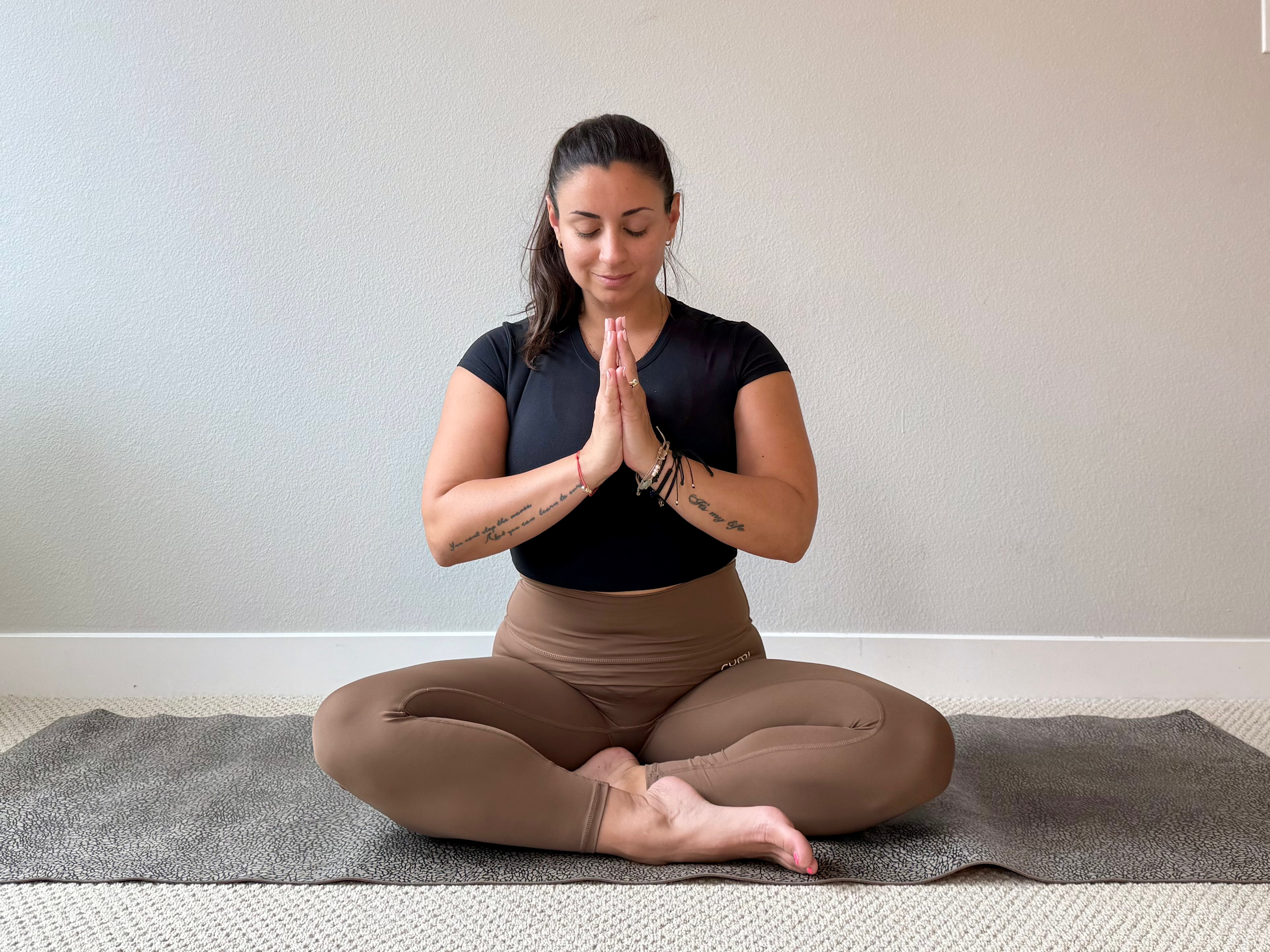 Elma Panagaki performs a cross-legged pose with her hands held together at her chest and eyes closed.