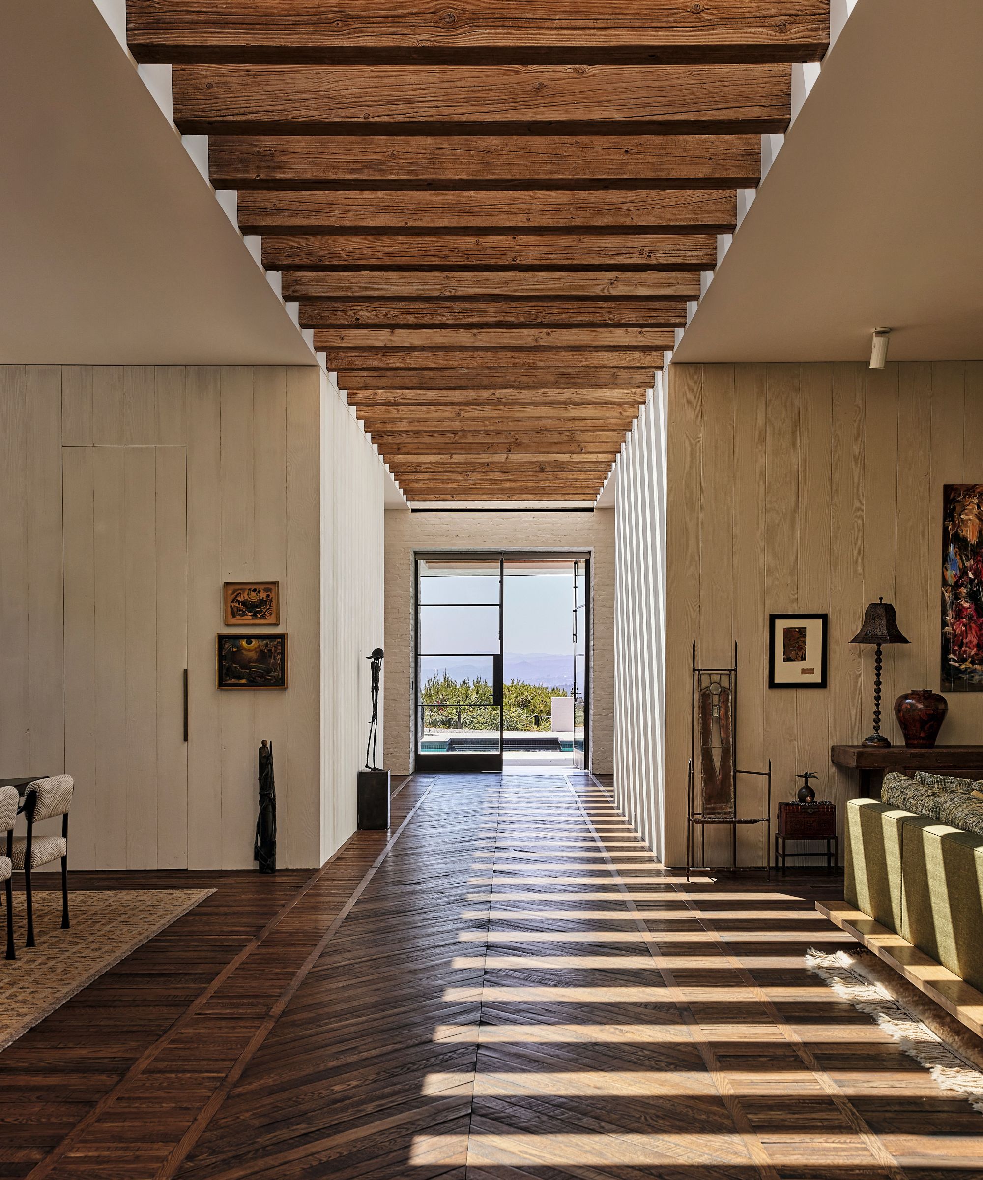 Sunlit hallway with timber-slatted ceiling, chevron wood floors, and linear light shadows leading toward a glass door opening to the outdoors