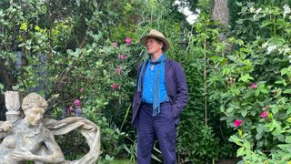 Monty wearing a cream hat with navy trousers and jacket standing in front of green foliage with a statue next to him