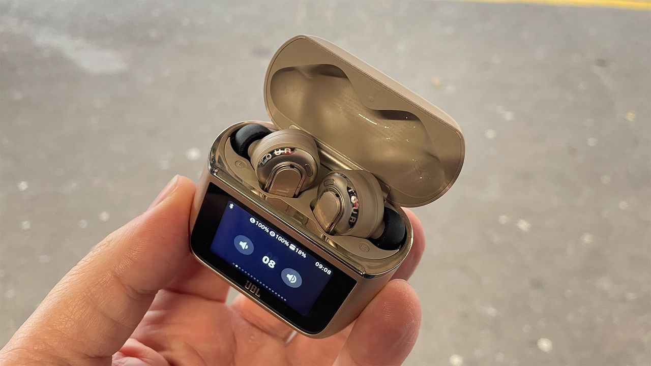 A hand holding the case for a pair of JBL Tour Pro 3 earbuds. The lid is open and the buds are visible inside. There are playback controls displayed on the screen on the front.