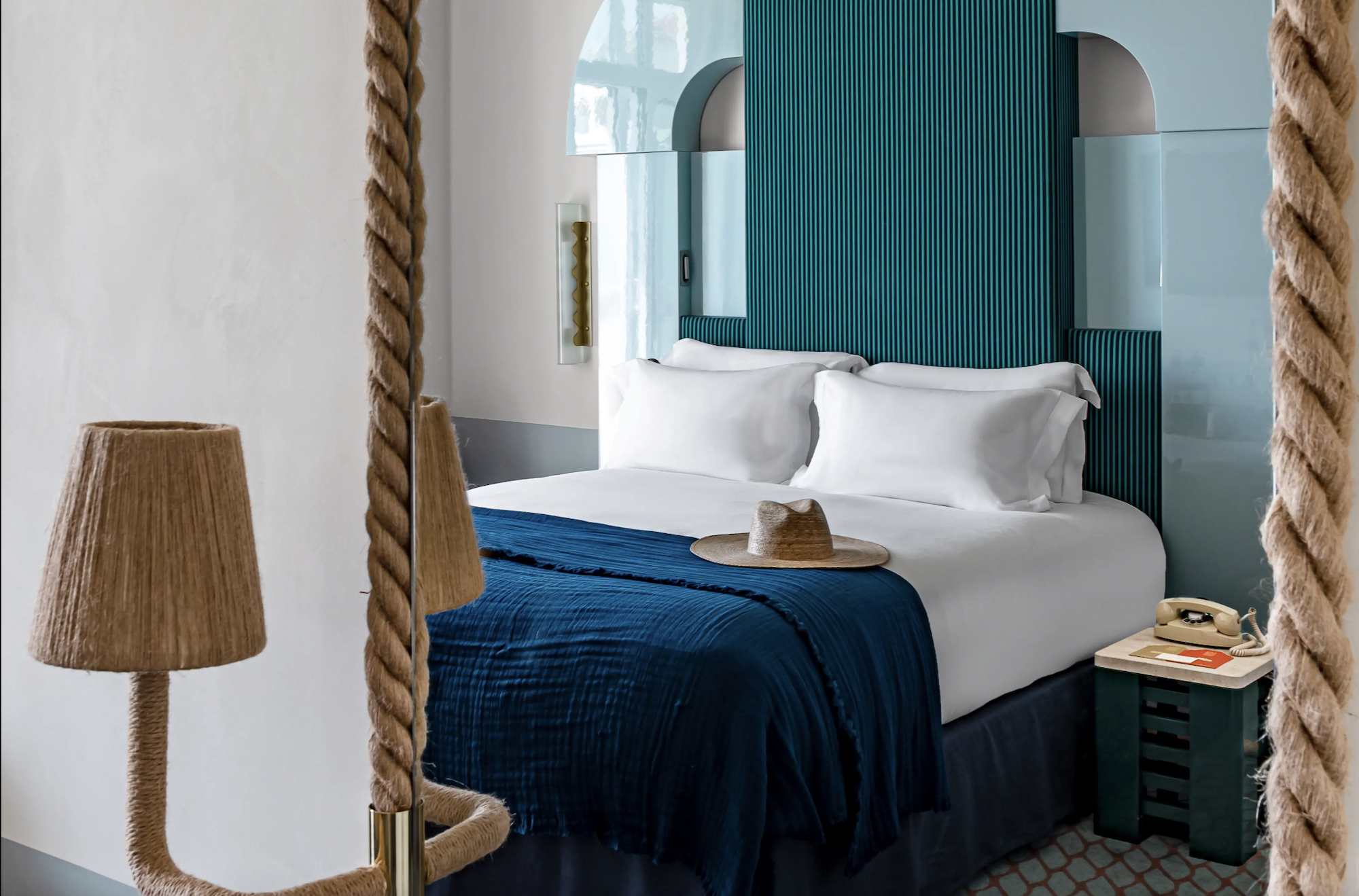 A view of a blue and teal coloured bed with a decorative, modern headboard and two nightstands either side. in the foreground is a rope framed mirror and a lamp.