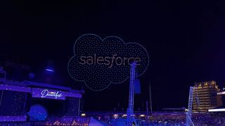 A swarm of drones equipped with LEDs form the Salesforce logo in the night sky, above the 'Dreamfest' closing party at Dreamforce 2024 in San Francisco. Decorative: The scenery below is lit in blue light, while the drones are lit in blue and orange.