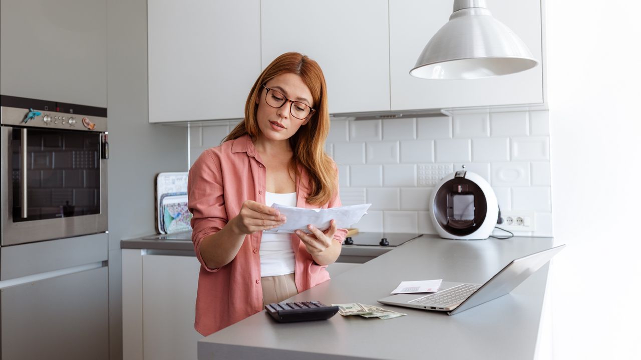 A woman looks at financial paperwork while standing in her kitchen, a calculator and laptop on the counter in front of her. 