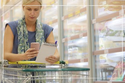 Woman in supermarket leaning on trolley and looking at her phone with a shopping list in hand