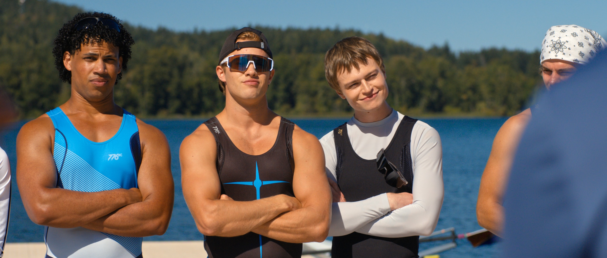 Three male athletes (l-r Jude Wilson as Baloo Garner, Sam Braun as Josh Regis, and Connor Paton as Ryder Regis) stand with their arms crossed in front of an unseen figure on a lake's dock, in Netflix's 'Crew Girl'.