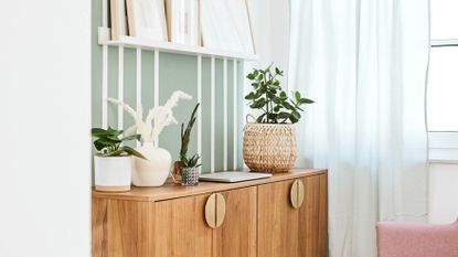 green room with a wooden side table with plants on top, and a white floating shelf full of artwork, with a white/green linen curtain against the window