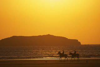 Two people riding horses are silhouetted against the sun on a beach in Morocco