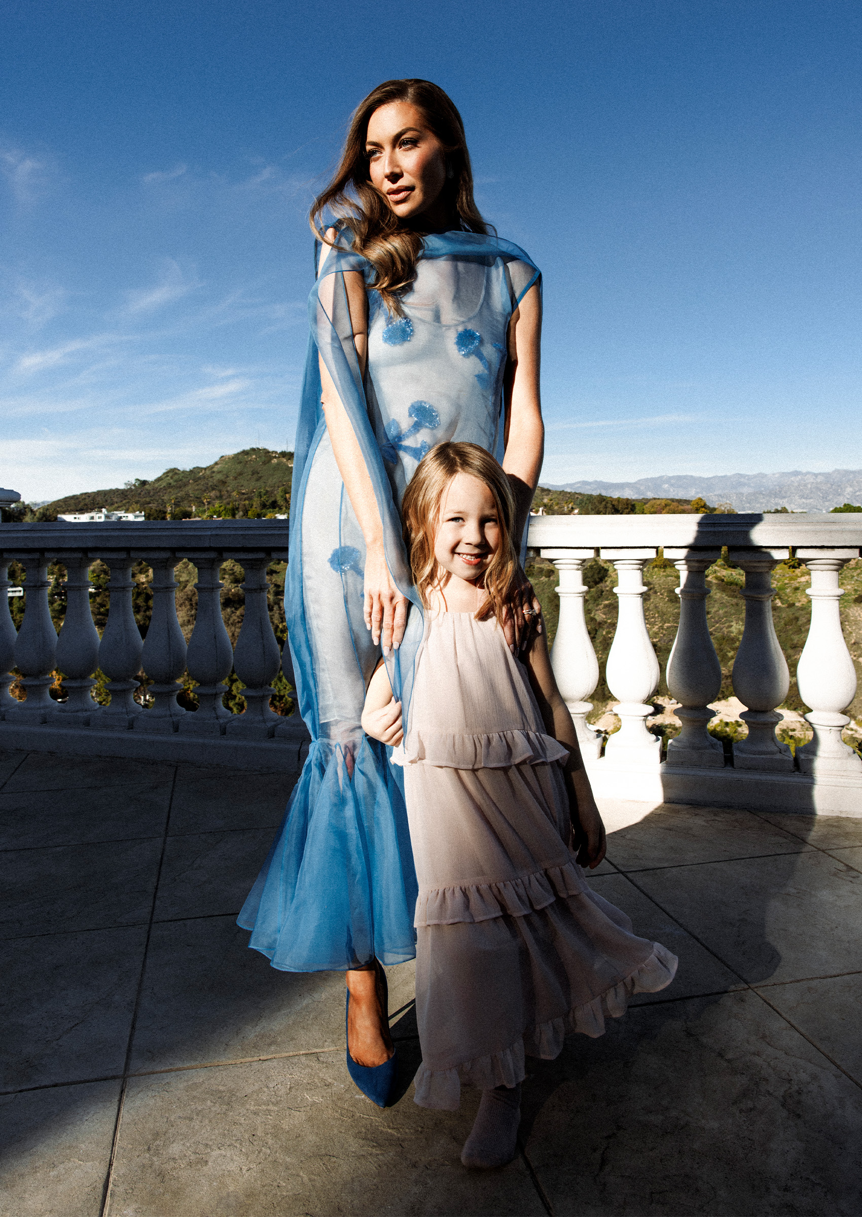 stassi schroeder wears a blue floral tulle gown and poses with her hands on her daughters shoulders on a balcony in los angeles