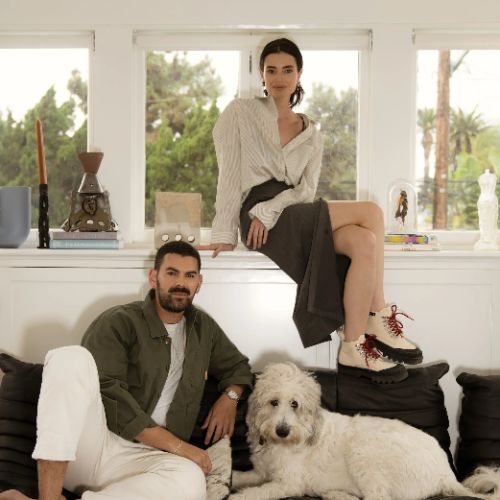 Image of a man sitting on a black sofa with a white dog next to him and a woman sitting on the windowsill above him.