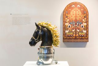 A horse's head (black with gold mane) on top of a silver object on wheels, with a closed, shuttered mirror on the wall behind