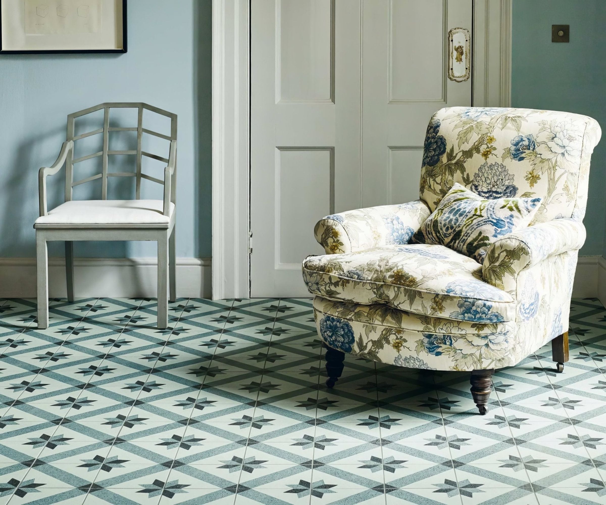 A charming room with pastel blue walls and patterned geometric floor tiles in soft green tones. A floral armchair and elegant painted chair add classic cottage style, highlighting how decorative tiles can elevate a traditional interior.