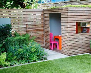 A wooden slatted fence and garden building behind a neat lawn and square flower bed in an urban garden.