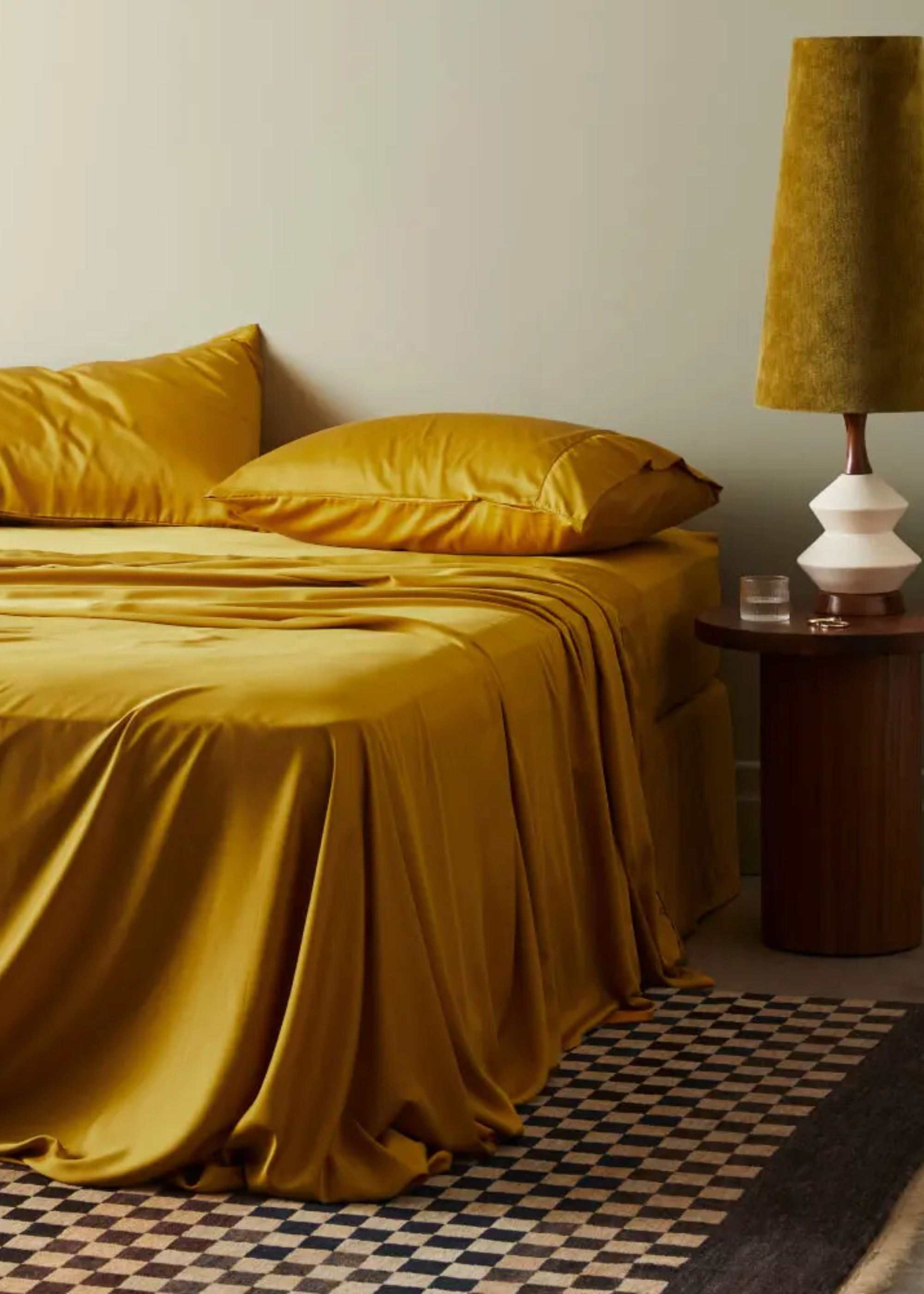 A bed with mustard yellow bamboo bedding, a checkerboard rug and a wood side table with a glass and a velvet lamp
