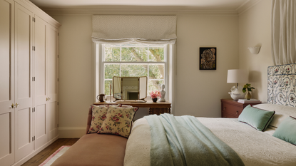 a bedroom with neutral colored walls, a wall of closets, a large window, a bed with cream colored sheets, a teal blanket, and teal pillows, and a bench at the foot of the bed with two floral pillows