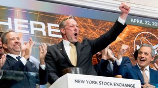 a man with dirty-blond hair and a light beard wearing a black suit, white shirt and tan tie pumps right fist in the air as he rings the closing bell at the new york stock exchange. Other men in suits applaud around him.