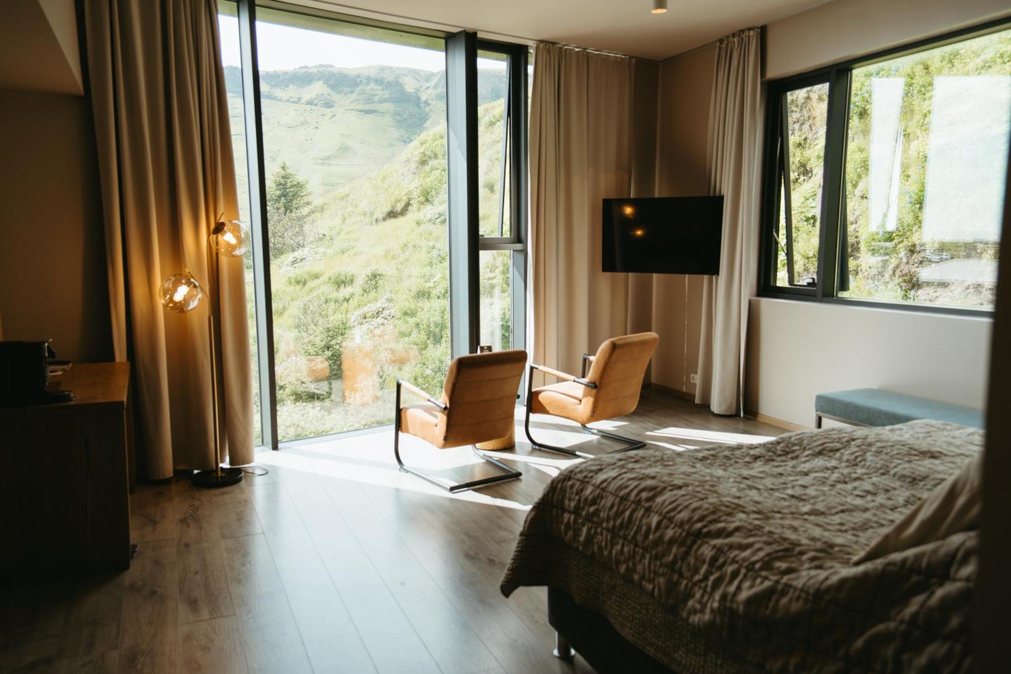 A mountain-view hotel room with two leather armchairs placed in front of a wide window, beige curtains, mellow light, and a checkered bed blanket.