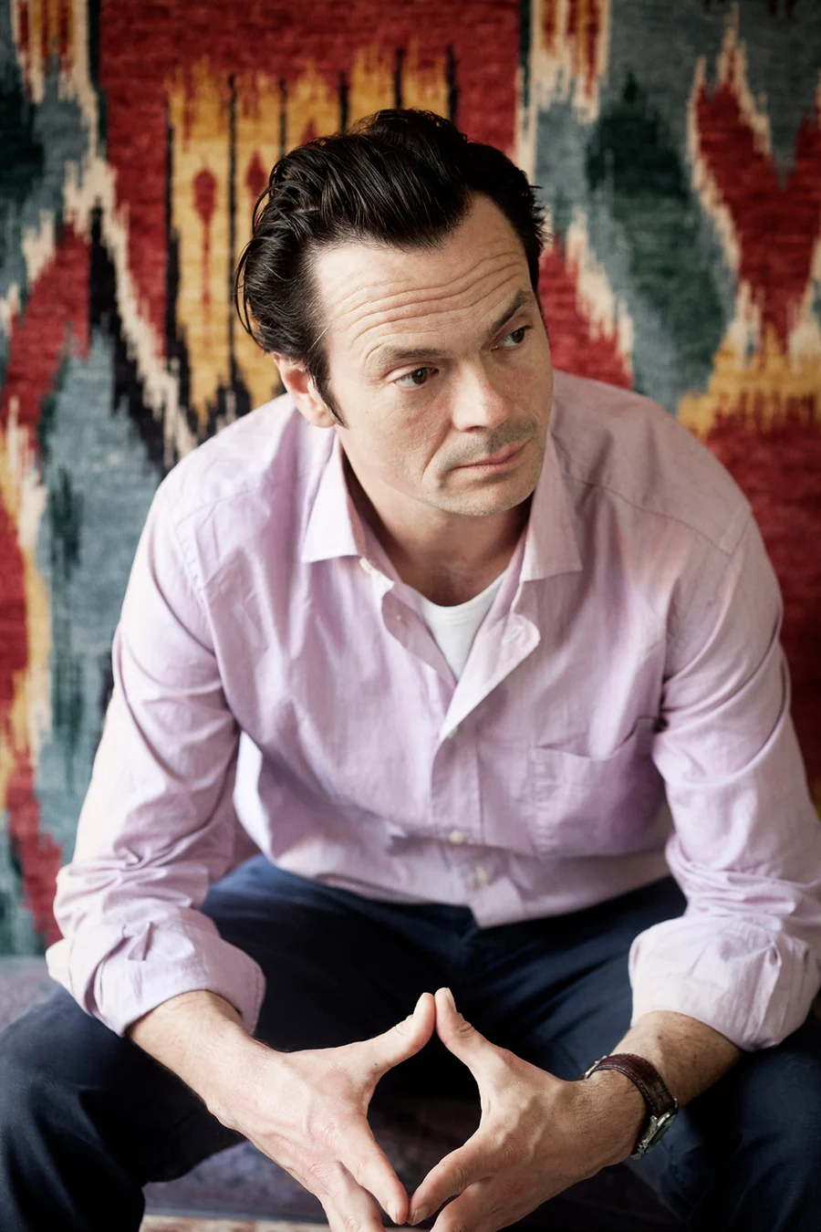 A man in a pink long-sleeved shirt and blue trousers looks to his left while posing sitting in front of a tufted red, blue, yellow, black, and white rug with his hands meeting.
