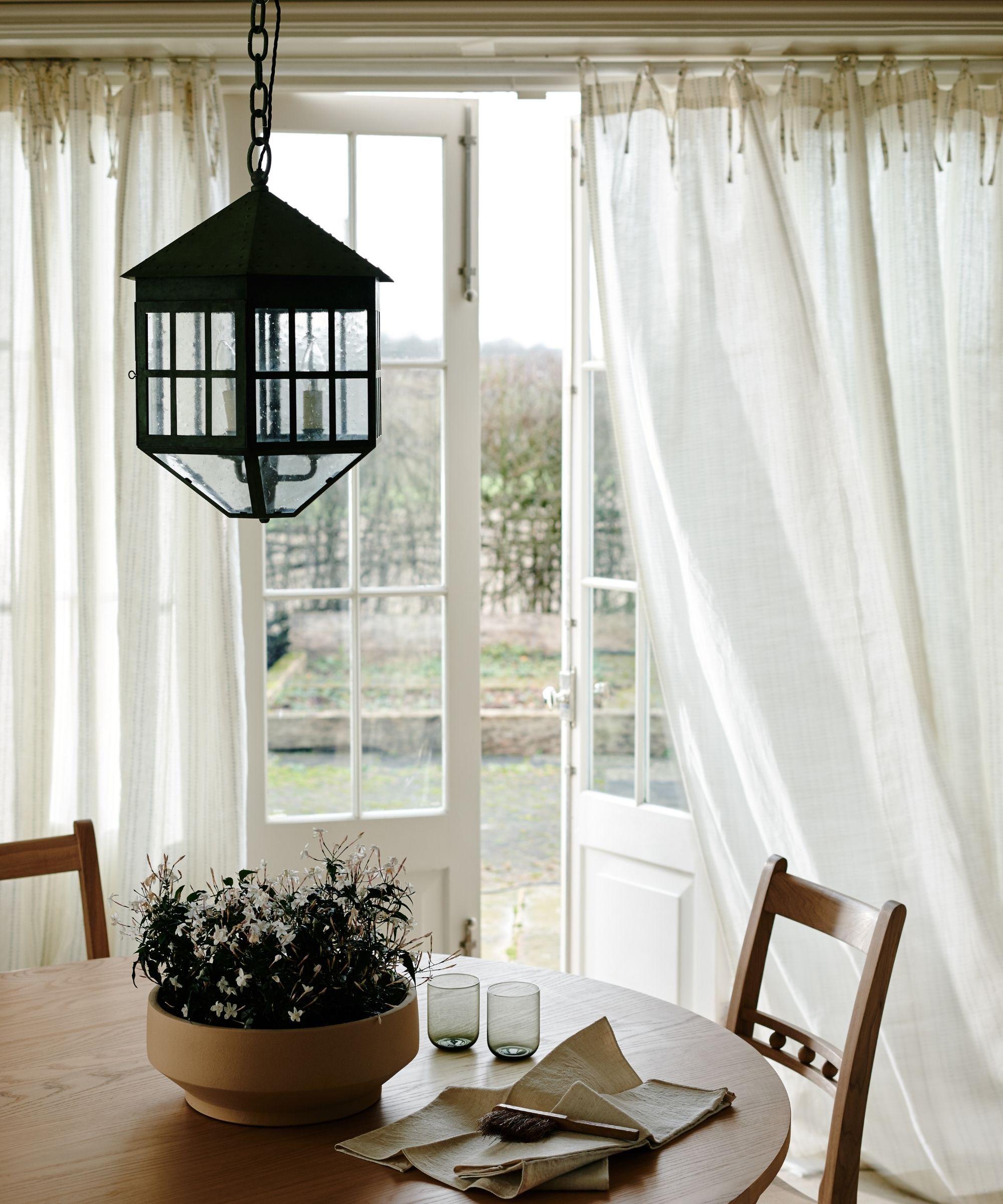 White open dining room doors with sheer white curtains and a circular wooden dining table and chairs.