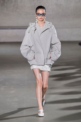 Model Tory Burch mengenakan jaket zip-up sporty abu-abu dengan rok mini, kacamata hitam, dan sepatu T-strap putih.