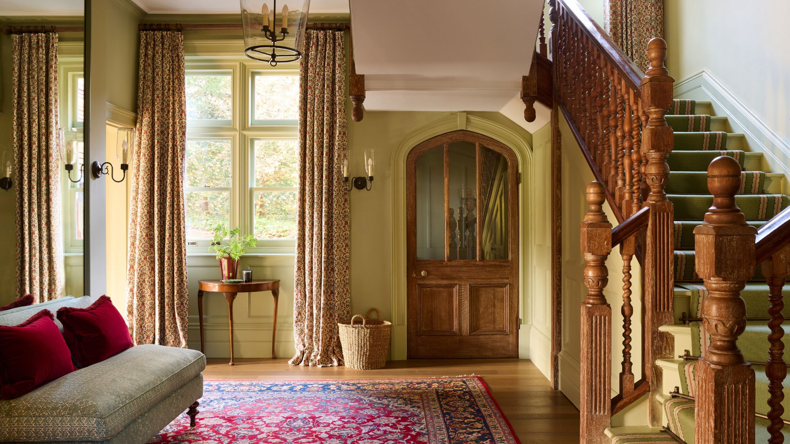 A large entryway in a traditional home with a curved staircase, light green walls, patterned curtains, wood floors with a raspberry red rug, and a small sofa with red cushions.