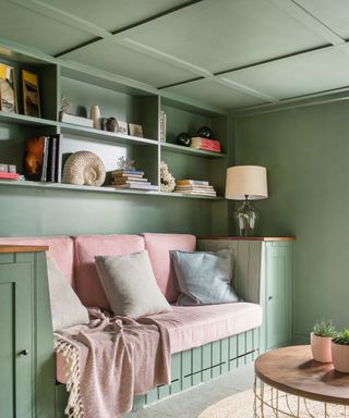 Seating space with painted green walls and ceiling, ceiling with paneling, seating area crafted from wooden paneling, finished with pink cushions and accessories, natural rounded rug and coffee table, shelves with decorative accessories and books