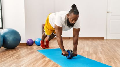 man wearing white tshirt yellow shorts and red trainers performing an abs rollout with an abs roller on a blue mat in a white gym space 