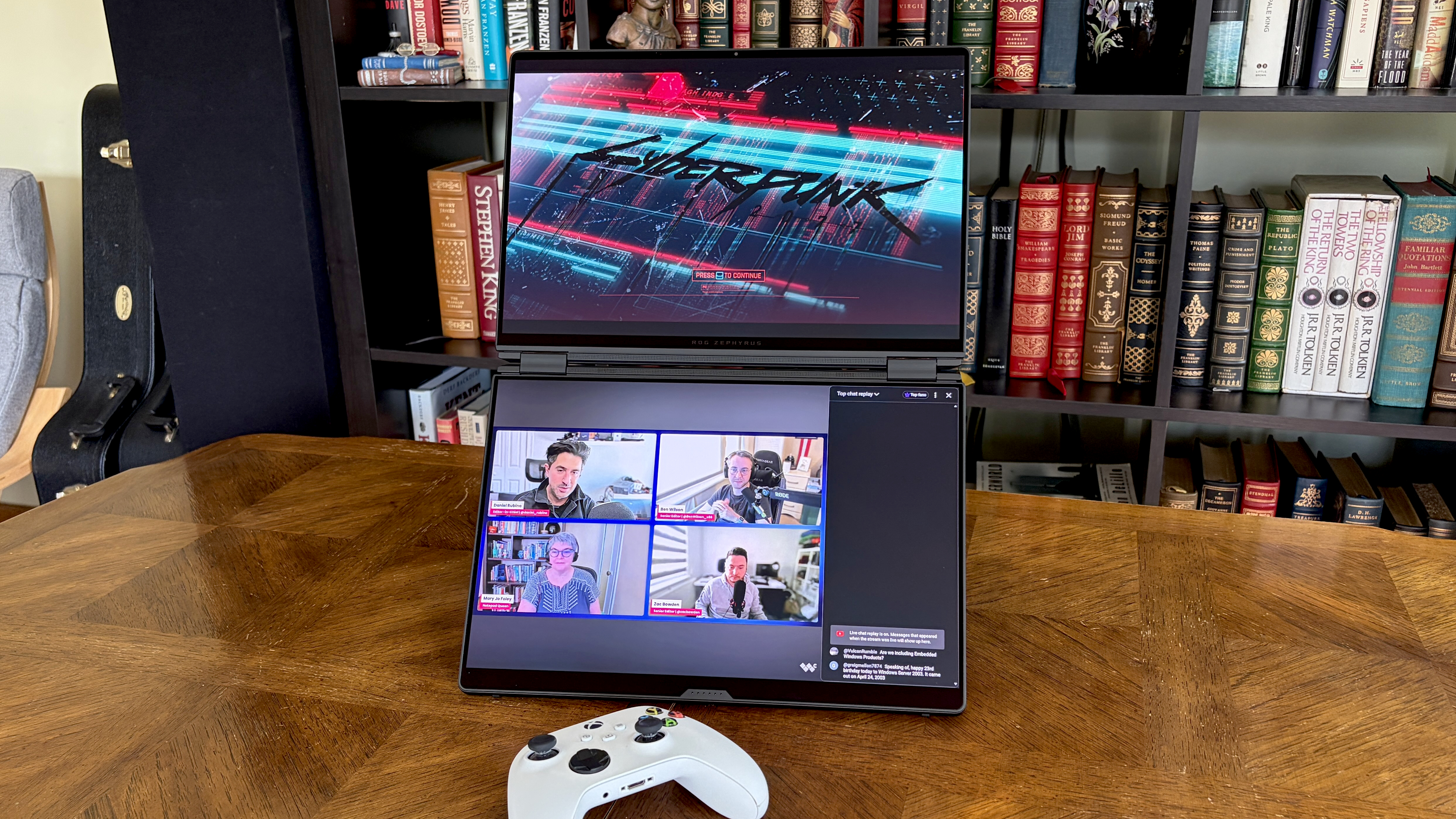 A gaming setup with two stacked monitors displays "Cyberpunk" on the top screen and a video conference on the bottom. A game controller rests on a wooden table, with bookshelves in the background.