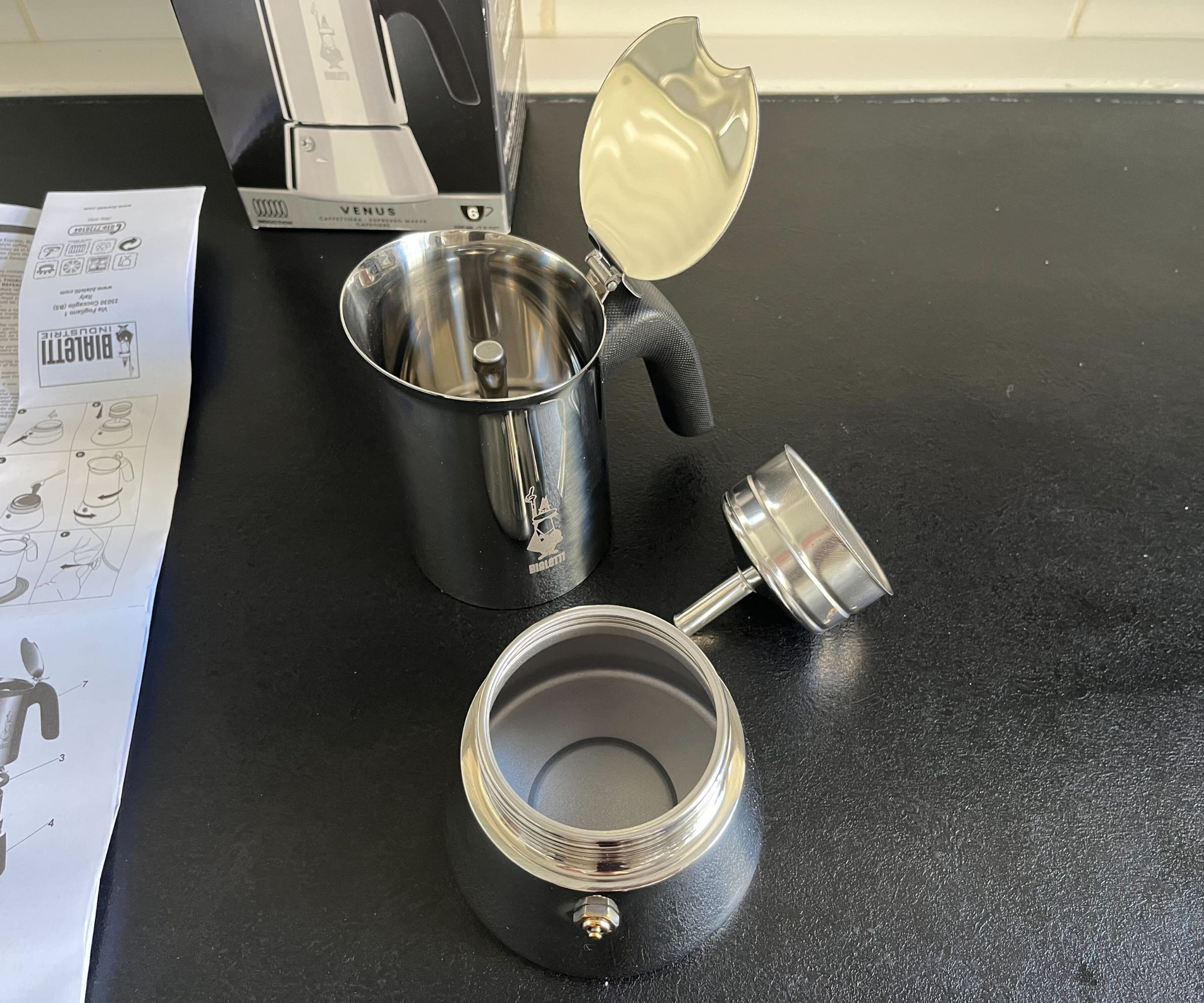 The dismantled Bialetti Venus coffee pot, with the base water trough, a metal filter, and the top jug section, on a black counter