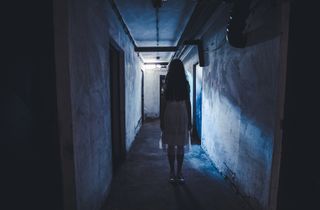 Dark-haired girl in white nightgown standing in concrete hallway with eerie light