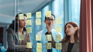 Office workers in a business strategy meeting discussing digital transformation plans with sticky notes placed on glass board.
